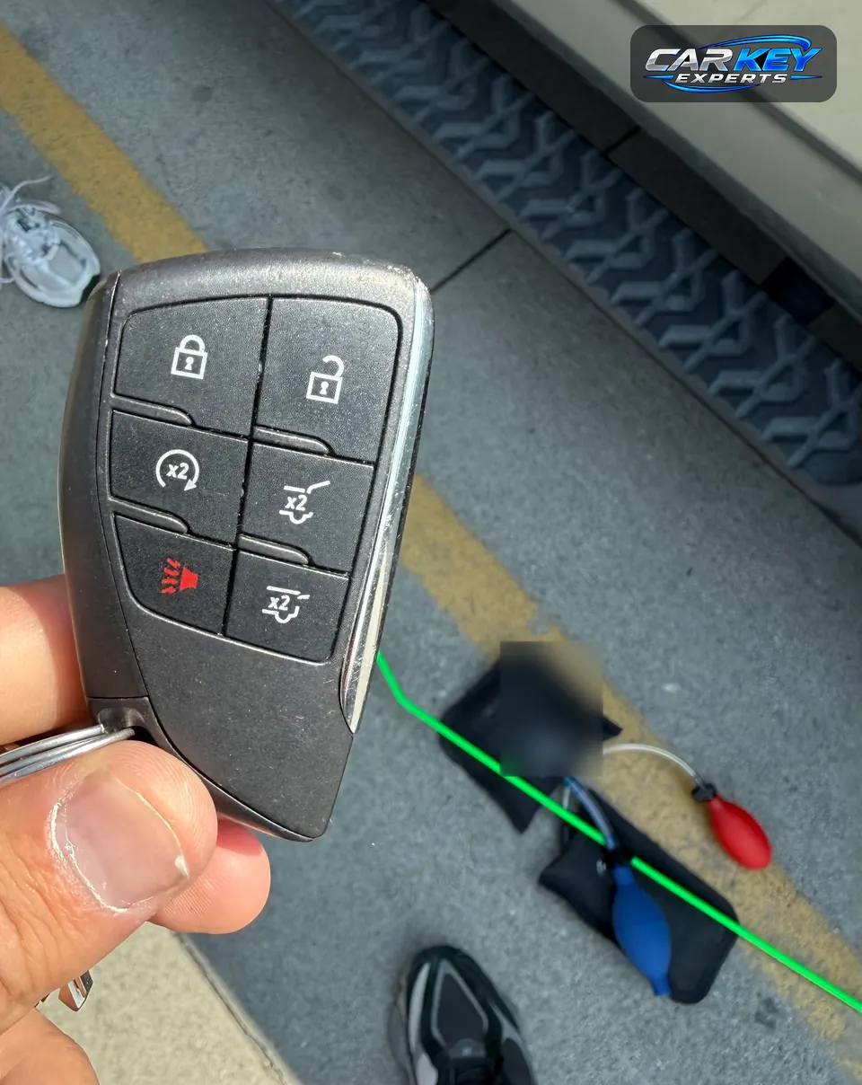 A six-button smart key held up over two air wedges and a long-reach tool lying on the pavement beside a vehicle