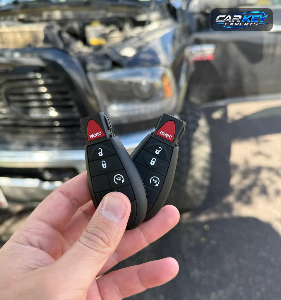 Two new fobik keys with remote start and panic buttons, held up in front of a pickup with its hood open