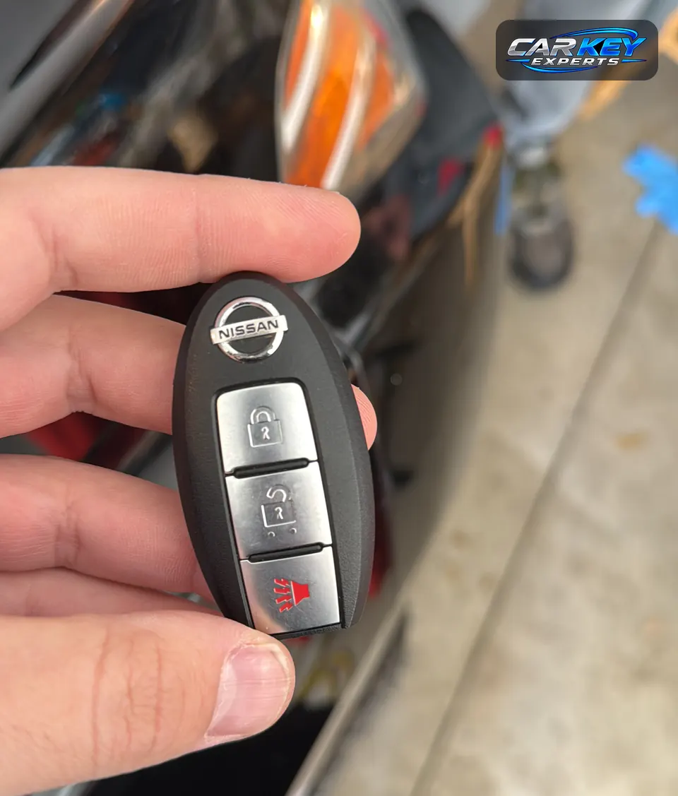 A Nissan smart key with lock, unlock and panic buttons, held up beside the headlight of a dark car