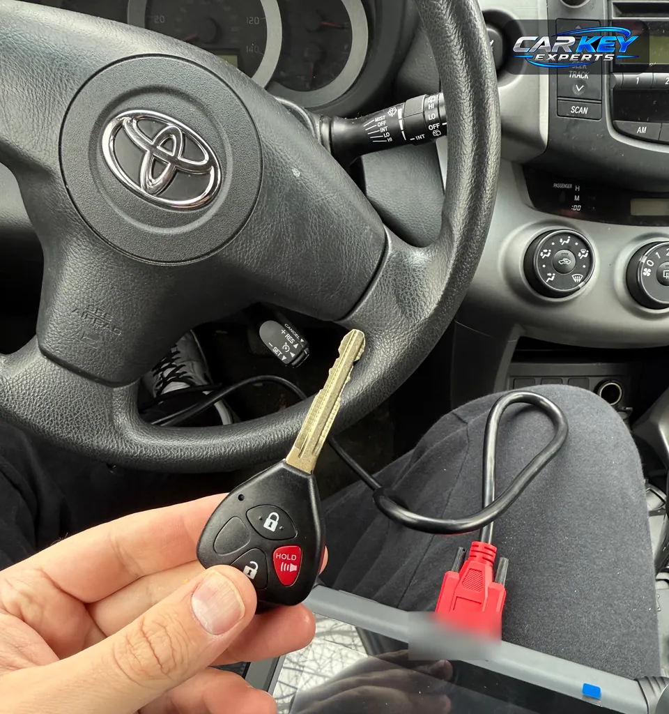 A Toyota remote-head key held up at the steering wheel, with the programming cable running to a tablet below, near Caldwell